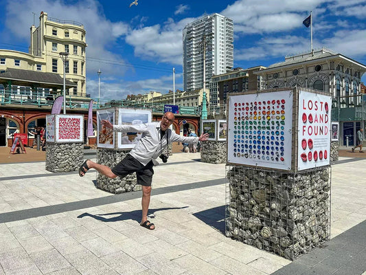 Photographer Alex Bamford playfully poses beside his Lost and Found exhibition on Brighton seafront. Display boards show colourful found objects and beachcombed items mounted on stone plinths, with Brighton buildings and the i360 area in the background.
