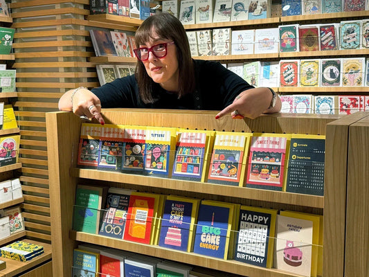 Lisa Bamford, creator of Stoats & Weasels and co-producer of the Brighton & Hove Calendar, pointing proudly at her colourful greeting cards on display in a shop.
