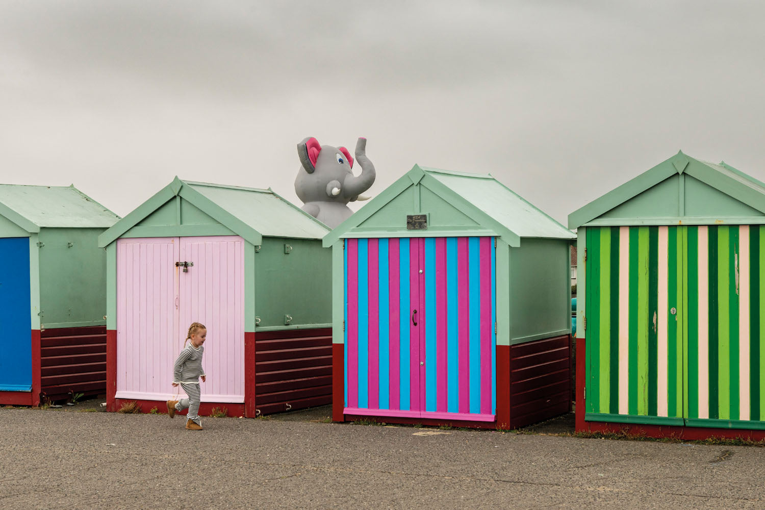 Brighton & Hove Calendar 2026 photo of a child running past colourful beach huts with a giant inflatable elephant peeking over the roofs