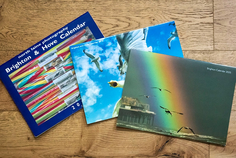Three past Brighton & Hove Calendars on a wooden floor, with colourful covers of seagulls, rainbows and beach scenes – part of the series now in its 2026 edition.