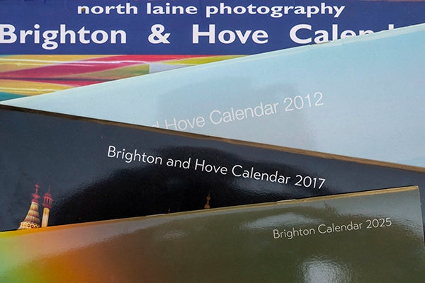 Stacked front covers of the Brighton and Hove Calendar from various years, including 2012, 2017, and 2025, showcasing different design styles and photography.