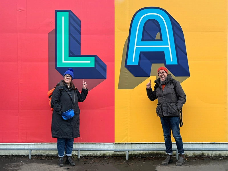 Lisa and Alex from the Brighton and Hove Calendar team standing in front of a bright mural featuring the letters "L" and "A", both smiling and pointing upwards.