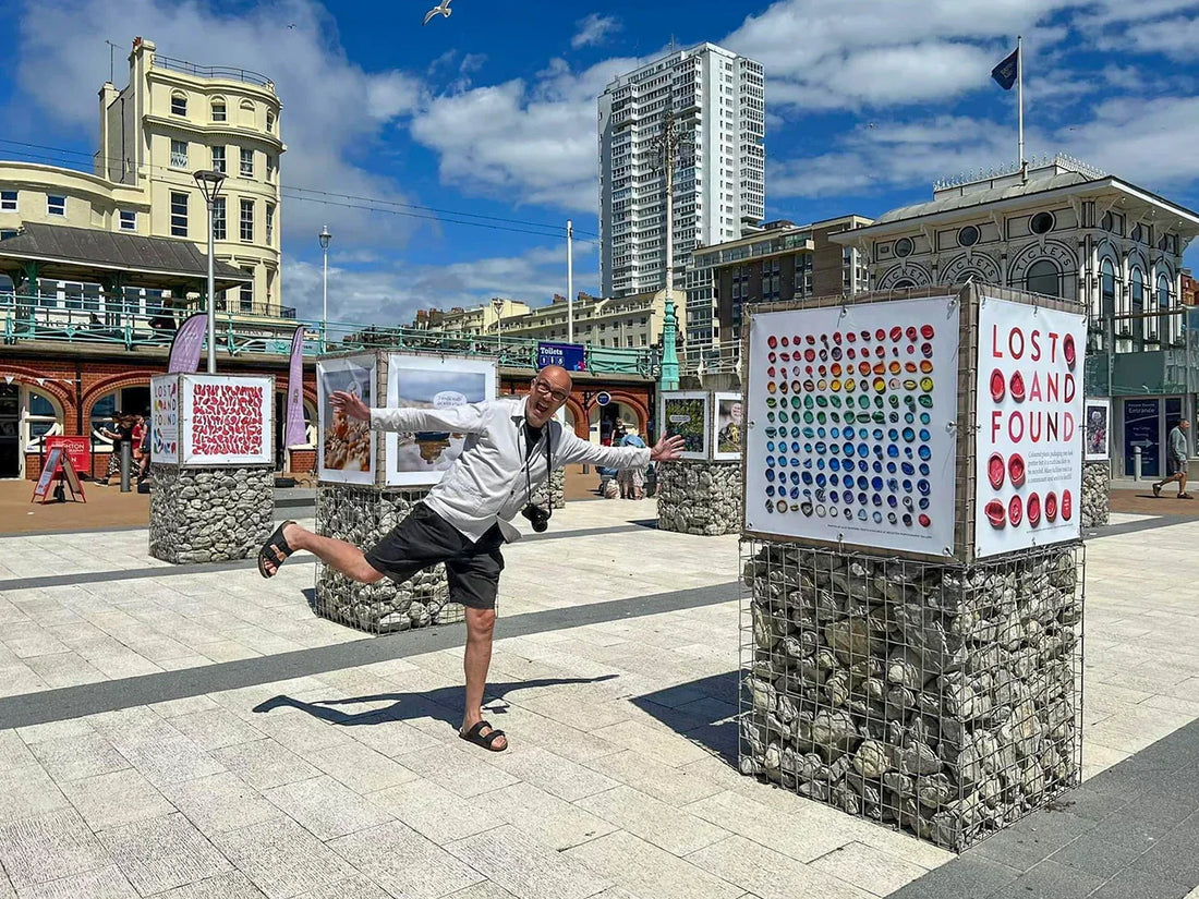 Photographer Alex Bamford playfully poses beside his Lost and Found exhibition on Brighton seafront. Display boards show colourful found objects and beachcombed items mounted on stone plinths, with Brighton buildings and the i360 area in the background.