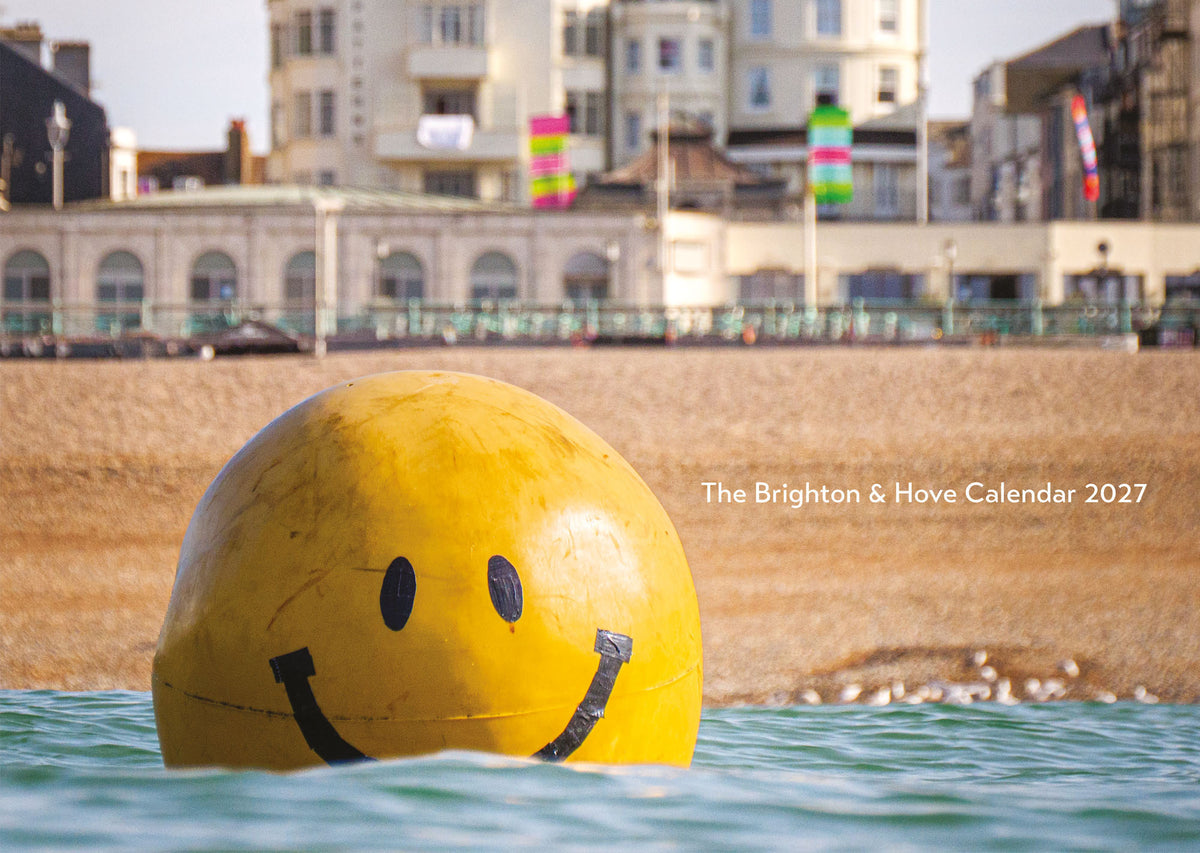 A large yellow smiley-face buoy floating in the sea at Brighton beach, with pebbles, buildings, and a clear sky in the background. White text on the right reads 'The Brighton & Hove Calendar 2027'.
