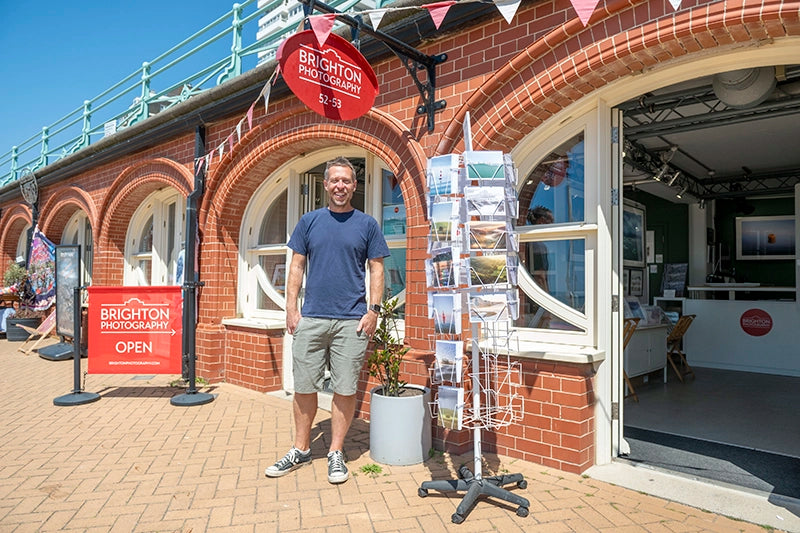 Finn Hopson stands outside Brighton Photography on the seafront, smiling beside a rack of postcards and a red sign that reads “Brighton Photography – Open”.