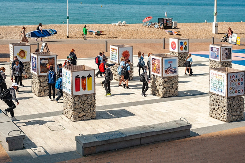 Visitors and school children exploring the 'Lost and Found' outdoor art exhibition on Brighton seafront, with the beach and sea in the background.
