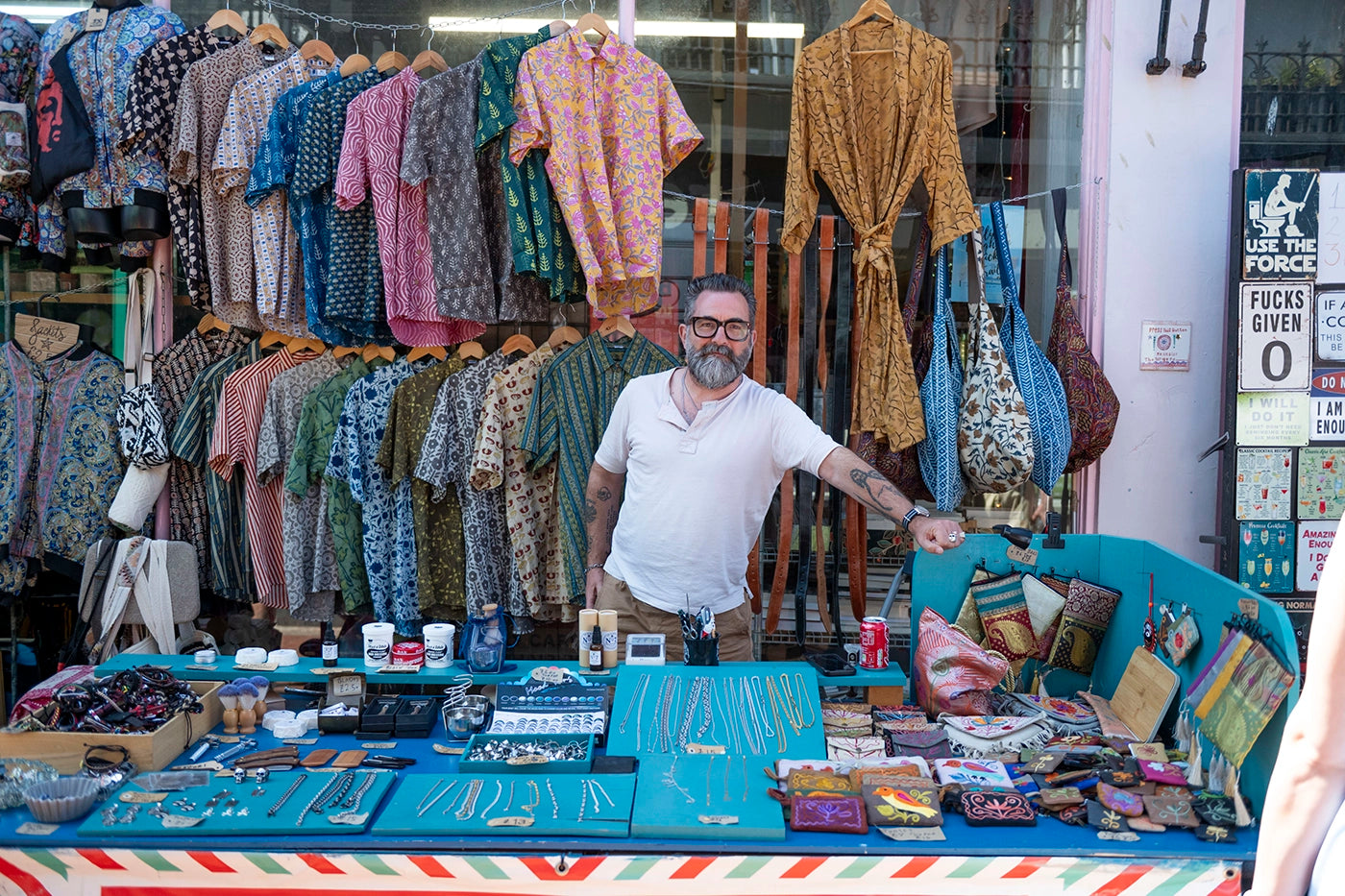 Marcus stands behind The Beard Man stall in Kensington Gardens, Brighton, surrounded by patterned shirts, jewellery, beard care products, and accessories. A colourful and eclectic market display set against a bright shopfront.